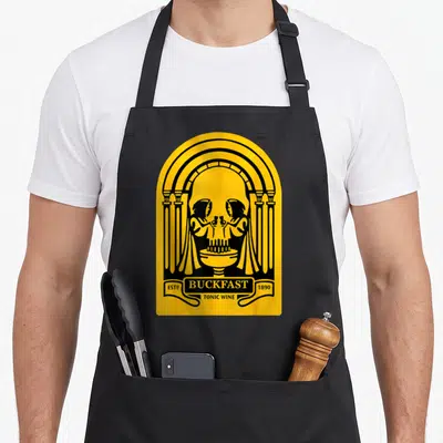 Buckfast wine apron - Yellow skull in black arch with two figures toasting in eye sockets and banner text — shown on a black cotton canvas apron against a white background
