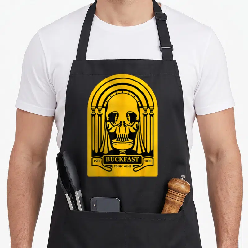 Buckfast wine apron - Yellow skull in black arch with two figures toasting in eye sockets and banner text — shown on a black cotton canvas apron against a white background