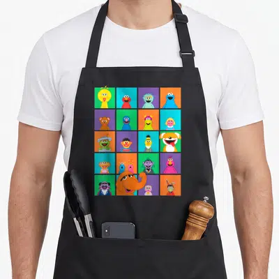 Square character aprons - Grid of muppet portraits in blue red yellow green purple squares — shown on a black cotton canvas apron against a white background