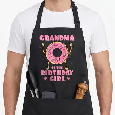 Doughnut grandma apron - Pink frosted donut character with GRANDMA OF THE BIRTHDAY GIRL text and two hearts — shown on a black cotton canvas apron against a white background