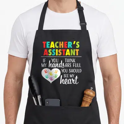 Assistant teacher apron - TEACHER'S ASSISTANT text with white heart containing red blue yellow green orange handprints — shown on a black cotton canvas apron against a white background
