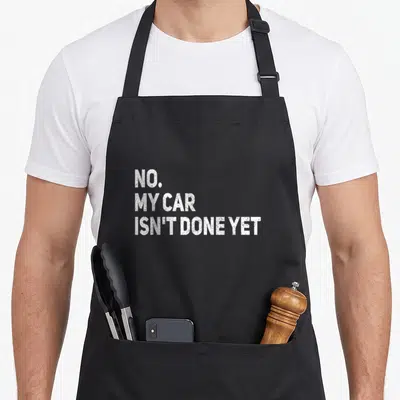 Ready car apron - White stacked text NO MY CAR ISN'T DONE YET on grey background — shown on a black cotton canvas apron against a white background