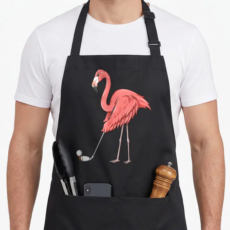 Golf flamingo apron - Cartoon pink flamingo holding silver golf club hitting white ball on brown tee — shown on a black cotton canvas apron against a white background