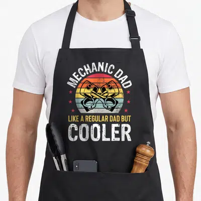 Car father auto mechanic apron - Crossed pistons over retro sunset stripes with MECHANIC DAD text — shown on a black cotton canvas apron against a white background