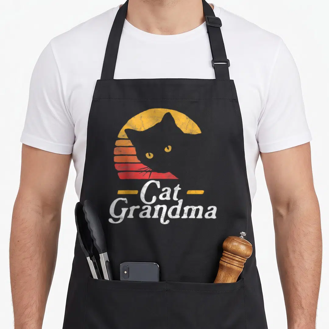 Cat grandma style aprons - Grey cat silhouette against yellow orange red striped sun with white text flanked by yellow — shown on a black cotton canvas apron against a white background