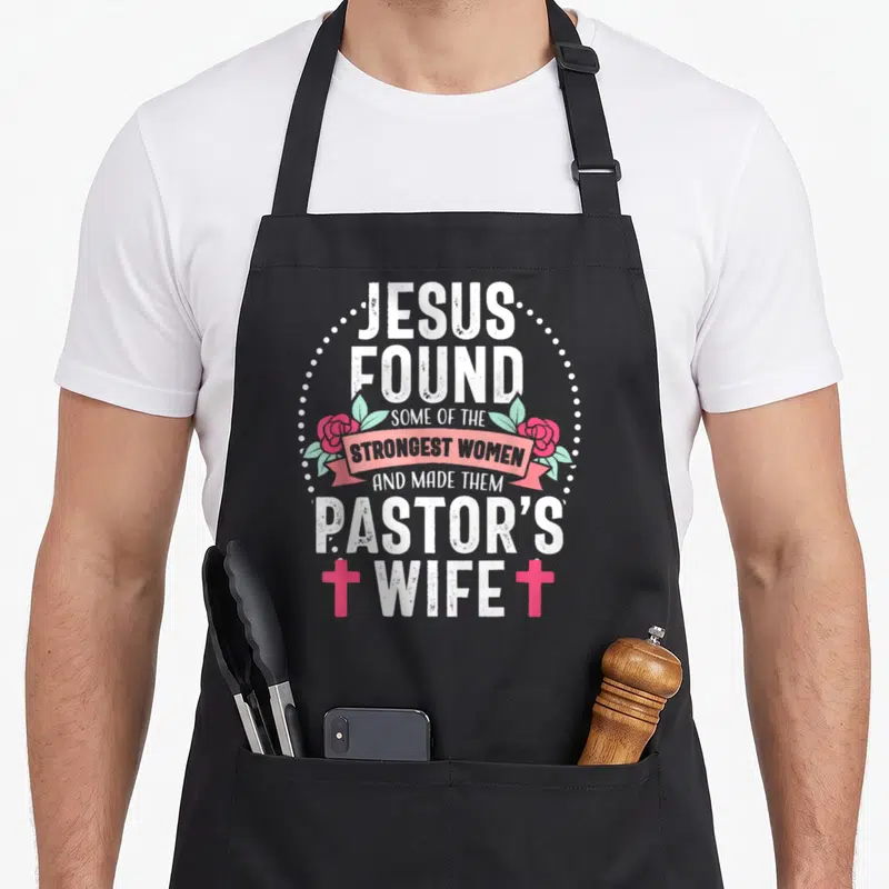 Jesus wife apron - Stacked white text pink ribbon roses two crosses dotted circle distressed — shown on a black cotton canvas apron against a white background
