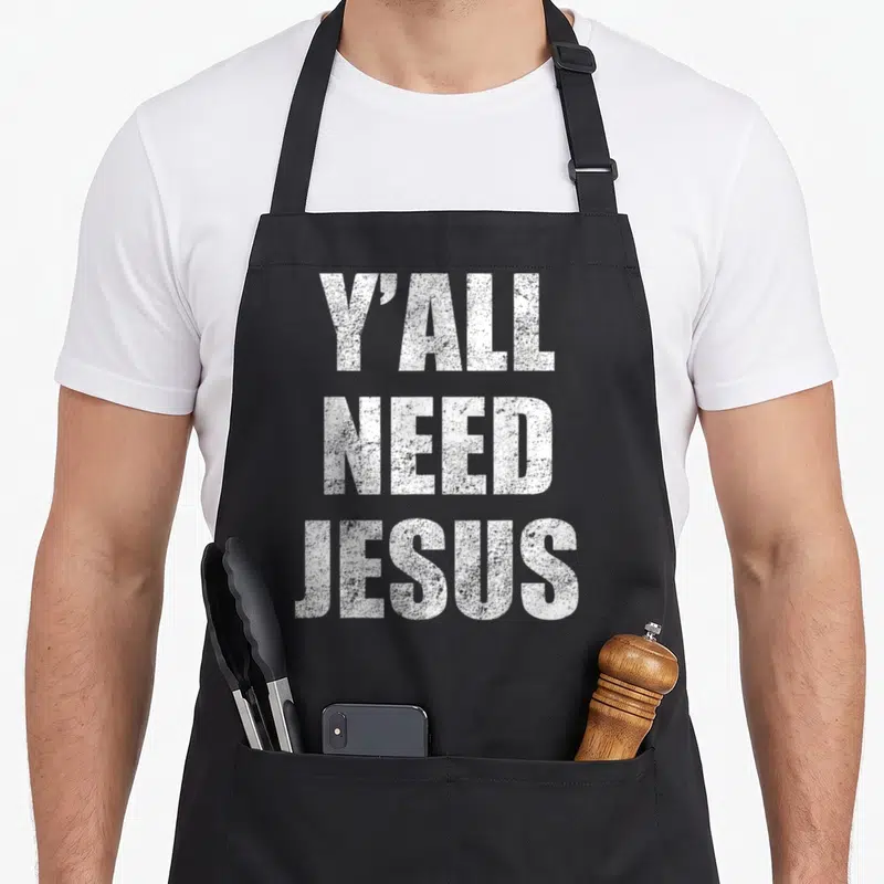 Yall christian apron - White Y'ALL NEED JESUS text stacked vertically on black background — shown on a black cotton canvas apron against a white background