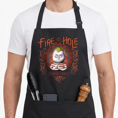 Pugsley hole apron - Pugsley Addams in red circle with Fire in the Hole text and spears — shown on a black cotton canvas apron against a white background