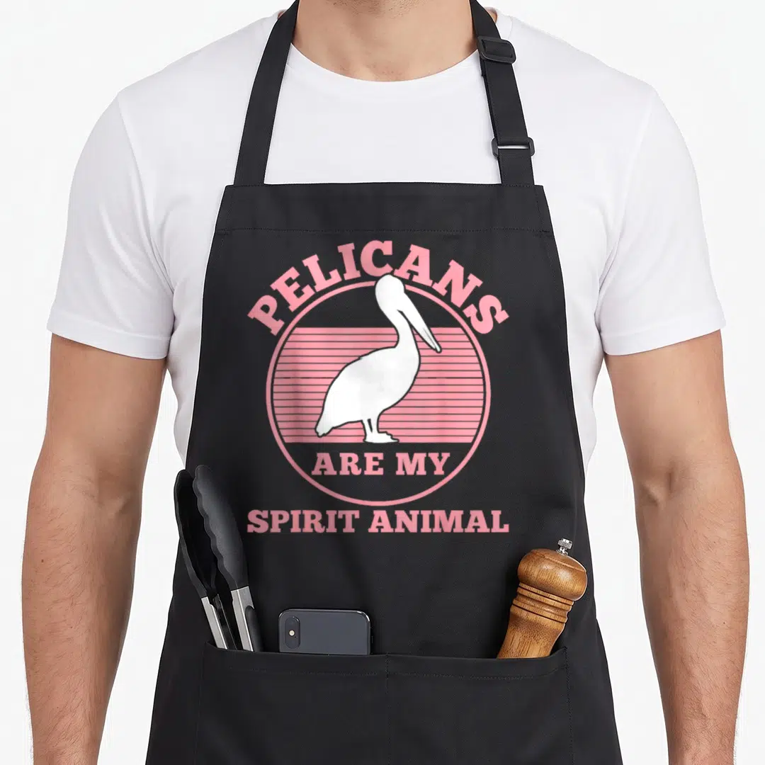 Cute pelican apron - White pelican inside pink striped circle with arched text above and straight text below — shown on a black cotton canvas apron against a white background