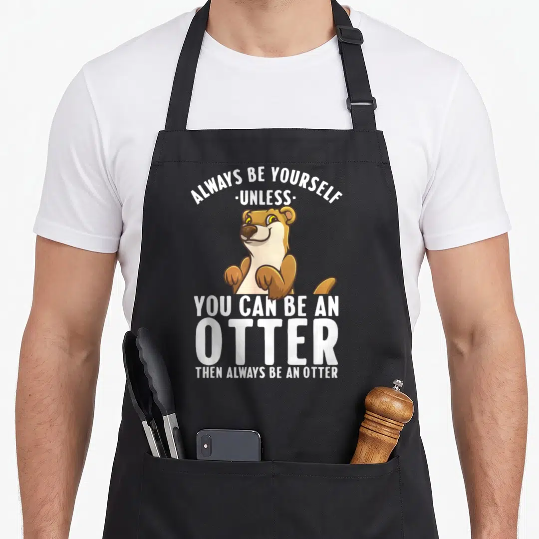 River animal apron - Cartoon otter in brown and tan with text Always Be Yourself Unless You Can Be An Otter — shown on a black cotton canvas apron against a white background