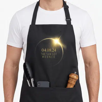 Accessory eclipse apron - Gold solar eclipse ring with date 04.08.24 and text THE SUN GOT MOONED centered inside — shown on a black cotton canvas apron against a white background