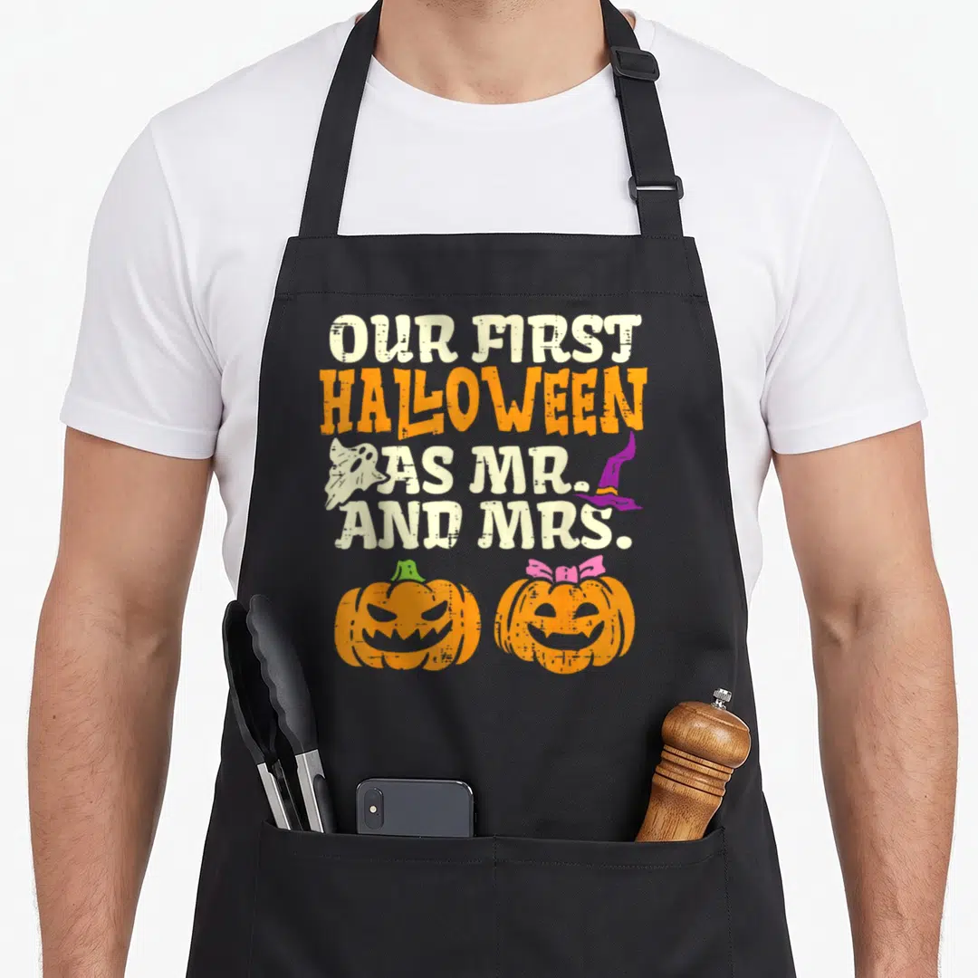 Our mr mrs aprons - Distressed text OUR FIRST HALLOWEEN AS MR AND MRS with ghost, witch hat, and two pumpkins, one — shown on a black cotton canvas apron against a white background