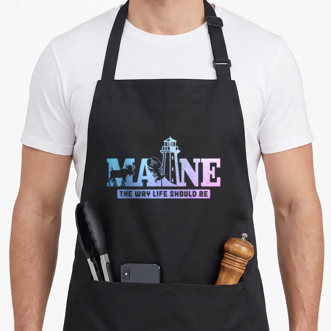 Lighthouse lobster apron - MAINE text with moose, lobster, and lighthouse in blue purple pink distressed style — shown on a black cotton canvas apron against a white background