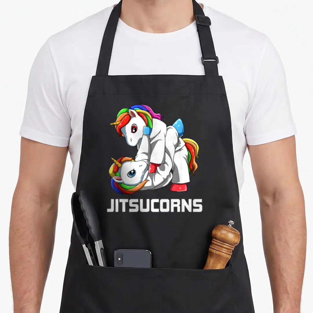 Bjj unicorn apron - Two unicorns in white gis grappling with JITSUCORNS text below — shown on a black cotton canvas apron against a white background