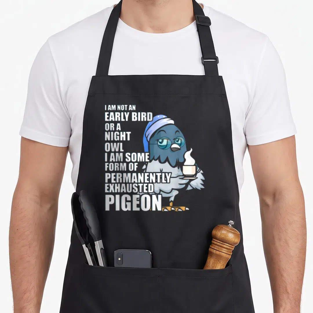 Permanently coffee apron - Cartoon pigeon holding steaming cup next to stacked white text about being permanently — shown on a black cotton canvas apron against a white background