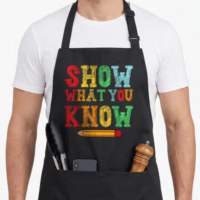 Exam testing teacher apron - Stacked red yellow green blue orange text reading SHOW WHAT YOU KNOW above a horizontal — shown on a black cotton canvas apron against a white background