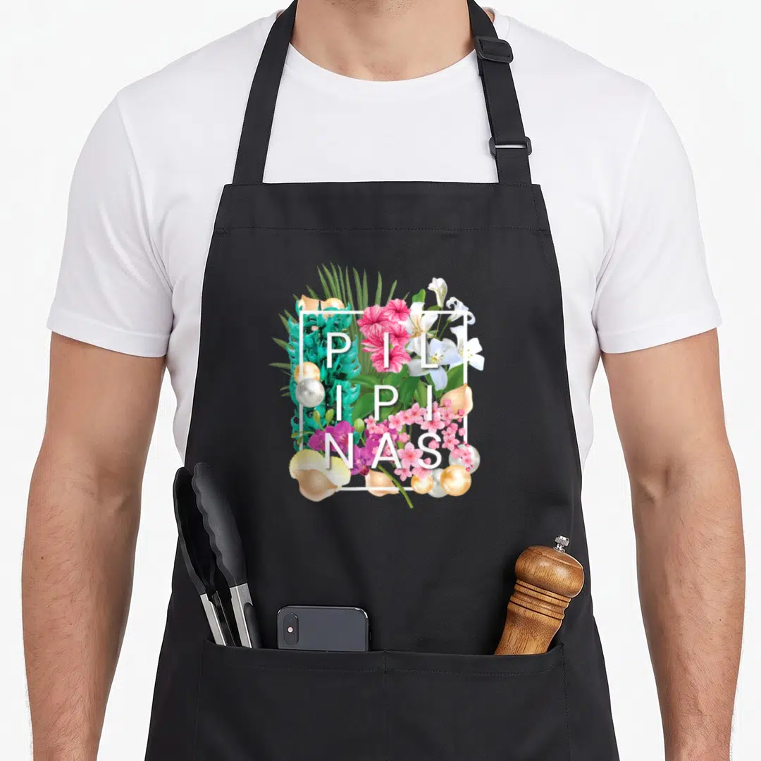Flower philippine pride apron - PILIPINAS text over pink white green gold flowers pearls shells in white square frame — shown on a black cotton canvas apron against a white background