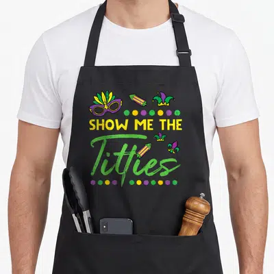 Show fat chef apron - SHOW ME THE Titties text with jester mask firecrackers and colored dots — shown on a black cotton canvas apron against a white background