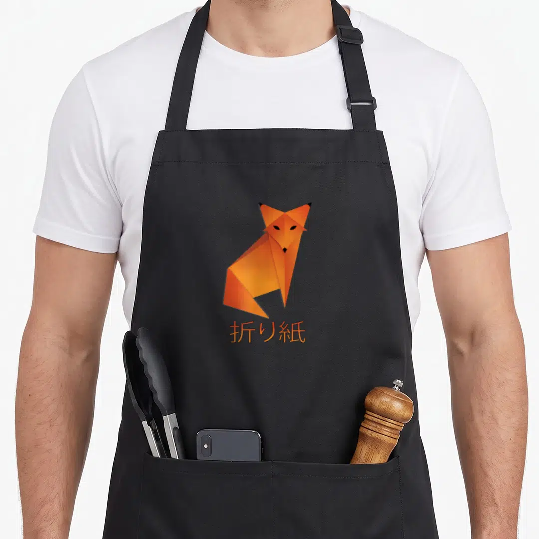 Origami fox aprons - Orange geometric paper fox above Japanese characters meaning origami — shown on a black cotton canvas apron against a white background