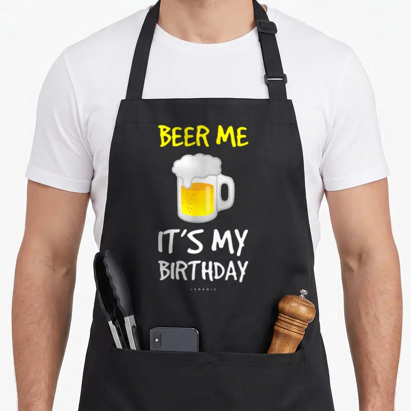 Birthday beer apron - BEER ME IT'S MY BIRTHDAY text surrounding a yellow cartoon beer mug — shown on a black cotton canvas apron against a white background