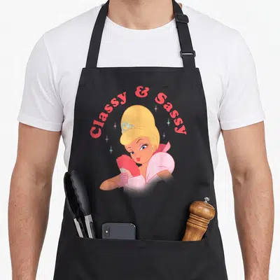 Charlotte princess apron - Cinderella holding a pink fan under arched Classy & Sassy text with sparkles — shown on a black cotton canvas apron against a white background