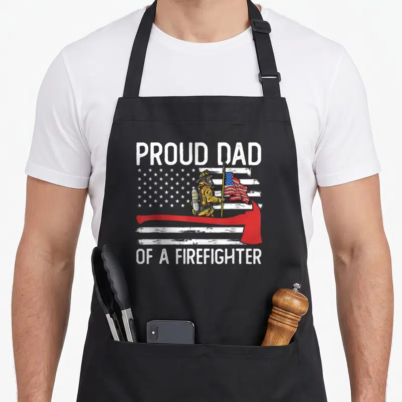 Fireman father firefighter aprons - Proud Dad of a Firefighter text with firefighter holding flag behind red axe — shown on a black cotton canvas apron against a white background