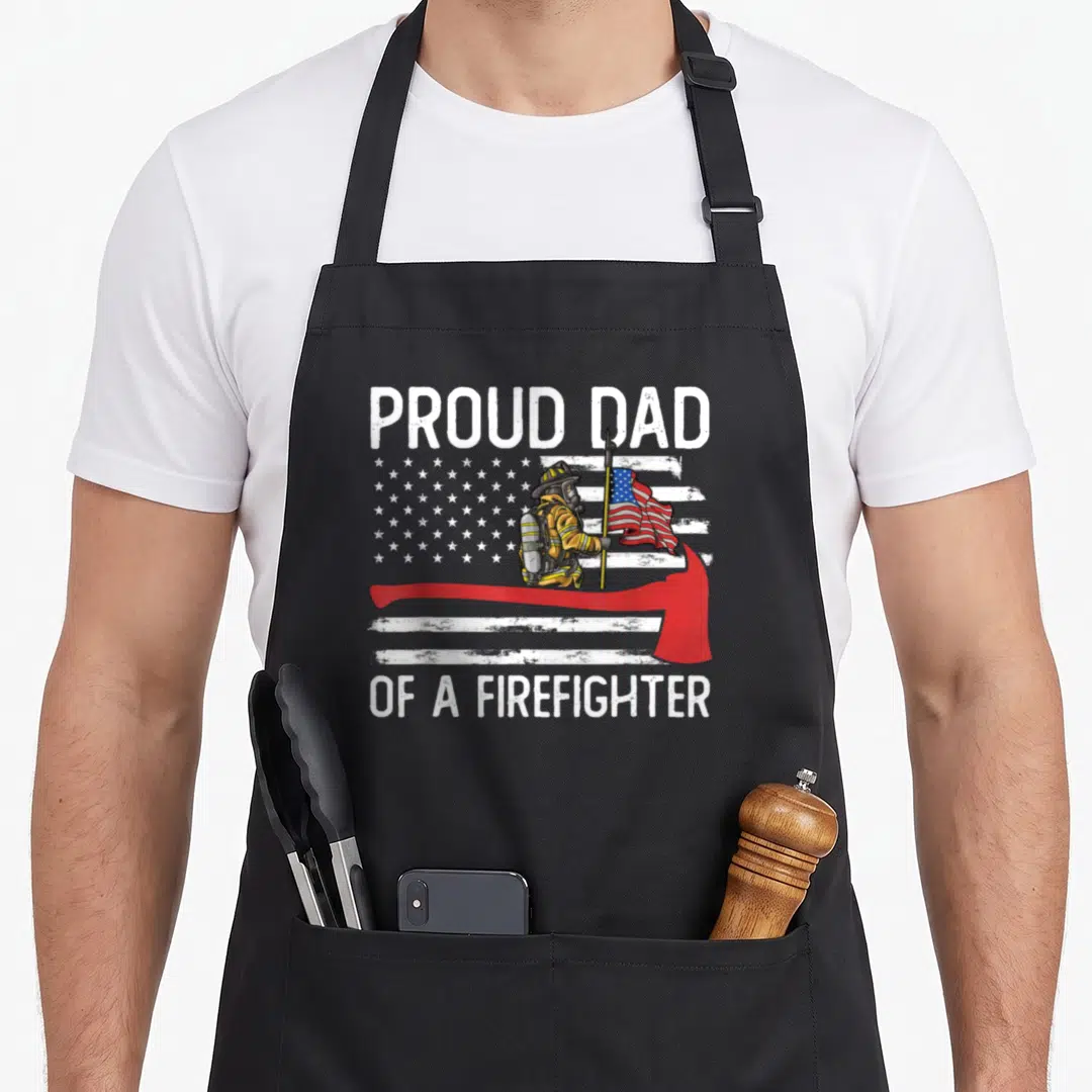 Fireman father firefighter aprons - Proud Dad of a Firefighter text with firefighter holding flag behind red axe — shown on a black cotton canvas apron against a white background