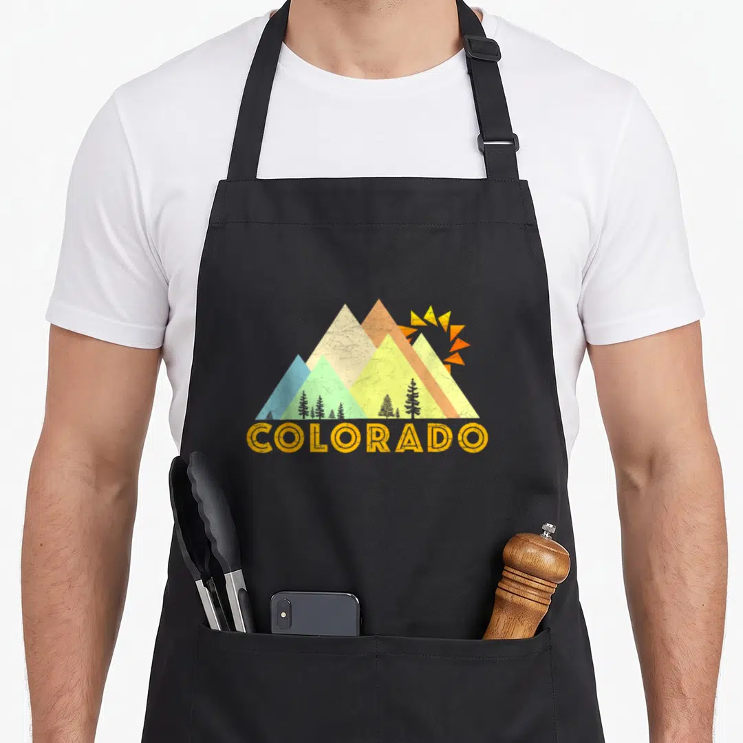 Funny distressed colorado apron - Retro mountain peaks with sun and pine trees above curved COLORADO text in orange — shown on a black cotton canvas apron against a white background