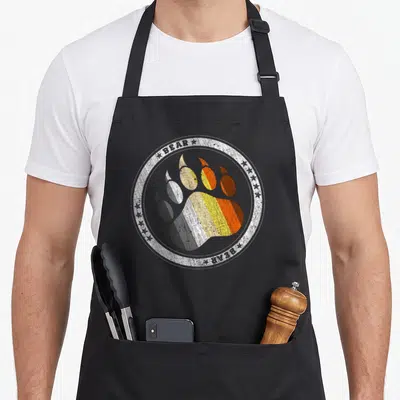 Paw gay pride apron - Bear paw print in brown orange yellow white and grey stripes inside distressed circle with text — shown on a black cotton canvas apron against a white background