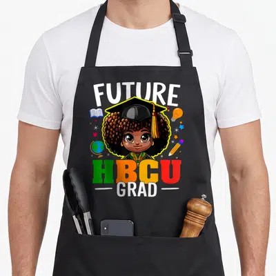 Grad future history apron - Cartoon girl in cap with FUTURE HBCU GRAD text and school supplies in yellow green orange — shown on a black cotton canvas apron against a white background