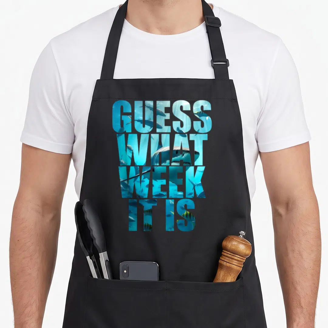 Guess summer aprons - Stacked text GUESS WHAT WEEK IT IS filled with blue and cyan sharks and fish — shown on a black cotton canvas apron against a white background