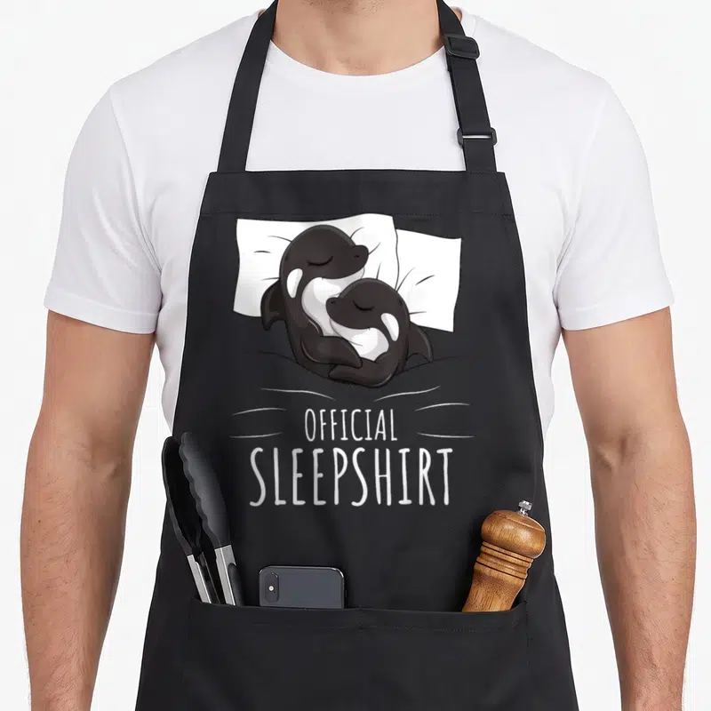 Orca novelty apron - Two cartoon orcas cuddling under a blanket with stacked text reading OFFICIAL SLEEPSHIRT flanked — shown on a black cotton canvas apron against a white background