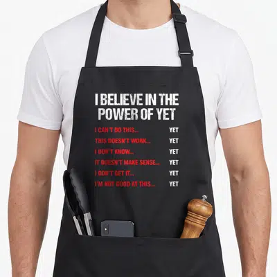 Encouragement judge dad apron - I BELIEVE IN THE POWER OF YET text in white and red stacked vertically with six red — shown on a black cotton canvas apron against a white background