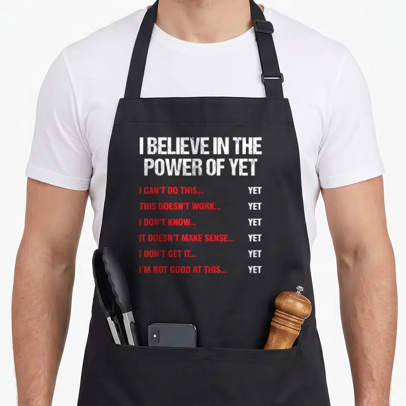 Encouragement judge dad apron - I BELIEVE IN THE POWER OF YET text in white and red stacked vertically with six red — shown on a black cotton canvas apron against a white background