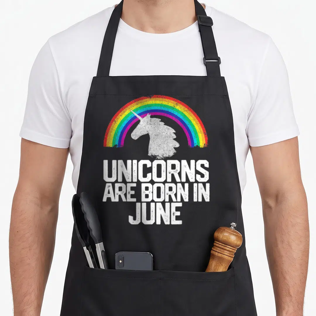 June rainbow apron - White unicorn head under rainbow arch with stacked text UNICORNS ARE BORN IN JUNE — shown on a black cotton canvas apron against a white background