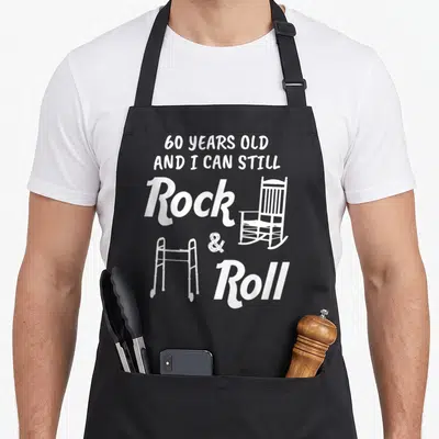 Gag 60th birthday apron - White text 60 years old with rocking chair and walker illustration — shown on a black cotton canvas apron against a white background