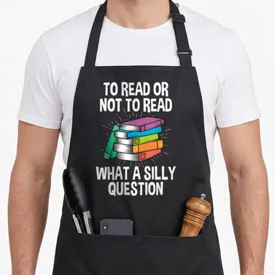 Read apron or apron - Four colorful books stacked with white rays and reading pun text in cartoon style — shown on a black cotton canvas apron against a white background