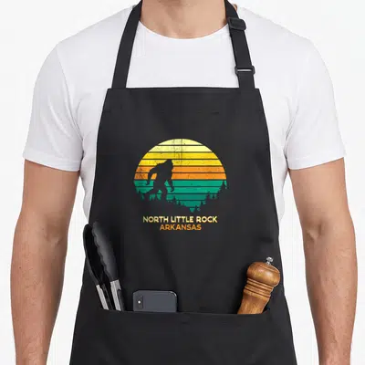 Arkansas bigfoot apron - Retro Bigfoot silhouette against striped sun with NORTH LITTLE ROCK ARKANSAS text — shown on a black cotton canvas apron against a white background