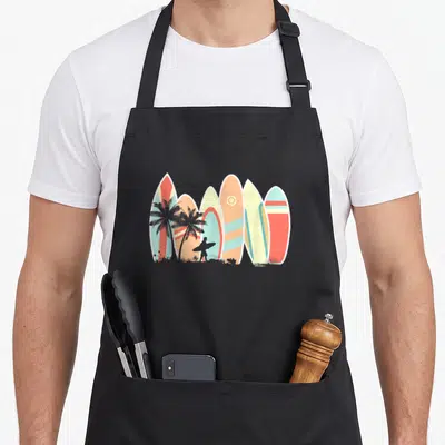 Surfboard surfer apron - Six colorful surfboards lined vertically with two palm trees and a surfer silhouette — shown on a black cotton canvas apron against a white background