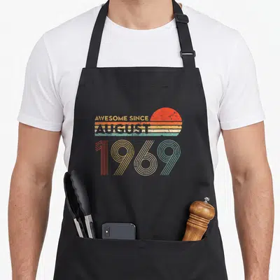 Funny august 50th birthday apron - Retro text AWESOME SINCE AUGUST 1969 with red sun and colored stripes — shown on a black cotton canvas apron against a white background