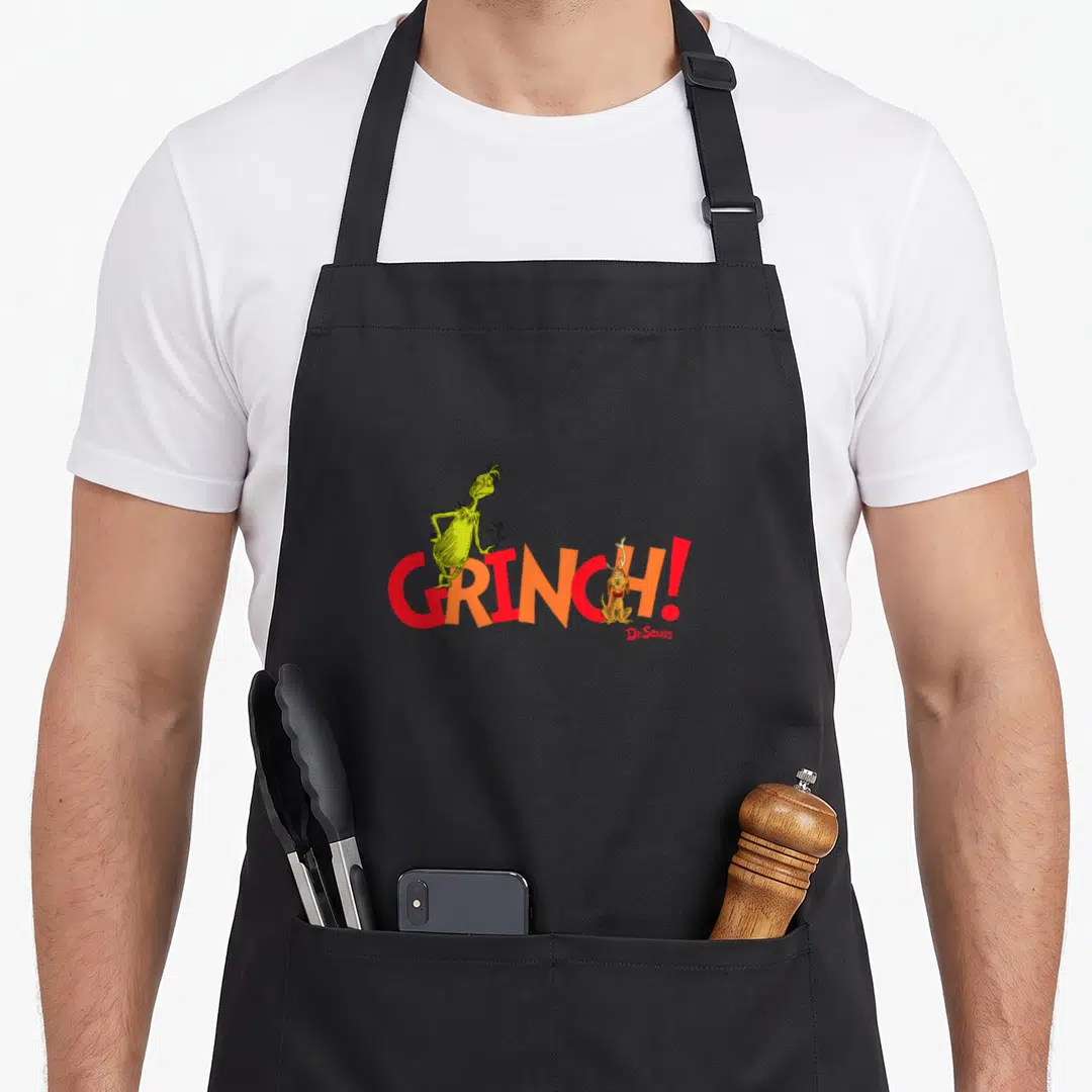 Cute max holiday apron - GRINCH text with lime green character and brown dog sitting right of exclamation point — shown on a black cotton canvas apron against a white background