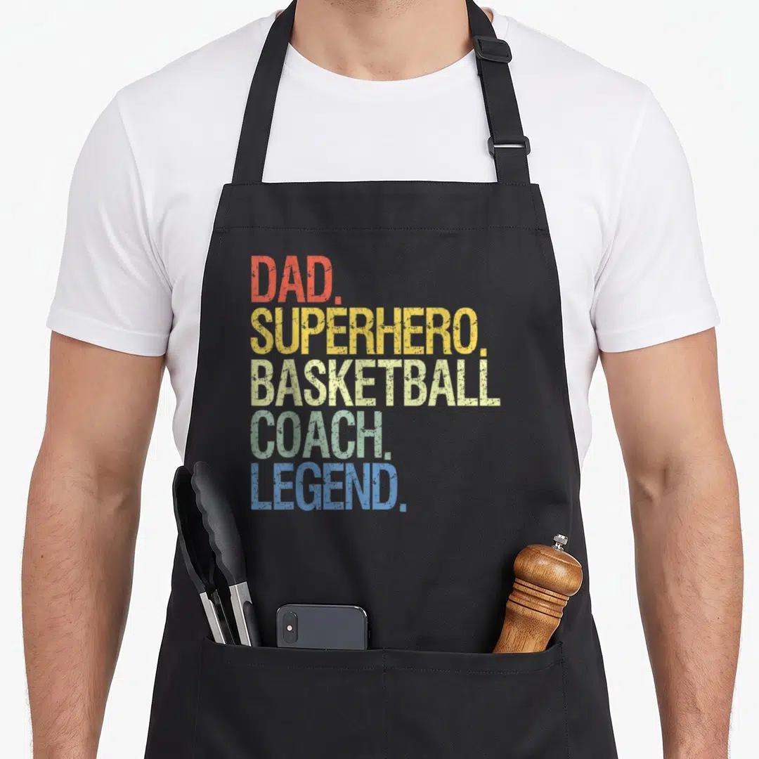 Basketball coach dad apron - Stacked distressed text DAD SUPERHERO BASKETBALL COACH LEGEND in red yellow green blue — shown on a black cotton canvas apron against a white background