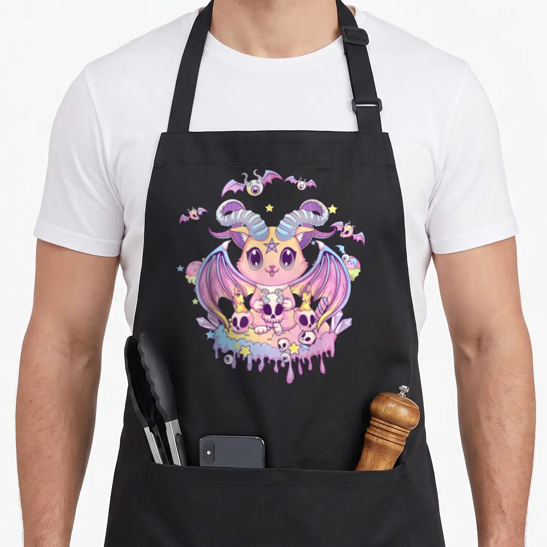 Krampus cat apron - Pastel demon cat holding skulls with bats, candles, crystals, and dripping slime in pink purple — shown on a black cotton canvas apron against a white background