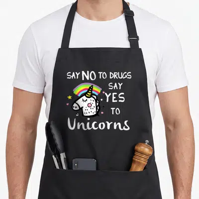 Ribbon unicorn apron - Cartoon unicorn head with rainbow and text Say No To Drugs Say Yes To Unicorns in white and black — shown on a black cotton canvas apron against a white background
