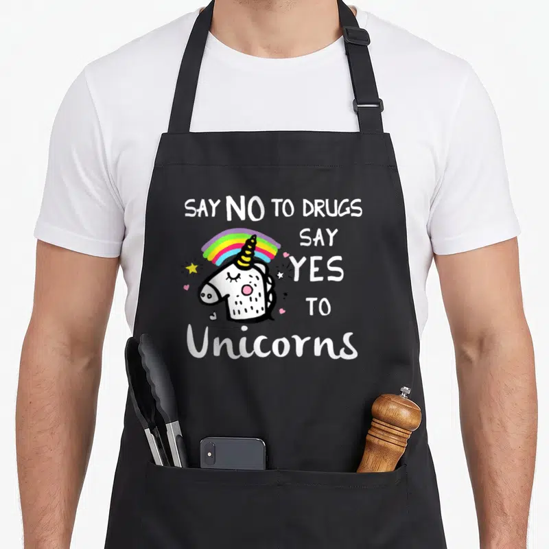 Ribbon unicorn apron - Cartoon unicorn head with rainbow and text Say No To Drugs Say Yes To Unicorns in white and black — shown on a black cotton canvas apron against a white background