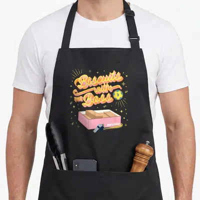 Funny biscuit boss apron - Biscuits with THE Boss text, pink box, whistle, blue cookie, yellow stars — shown on a black cotton canvas apron against a white background