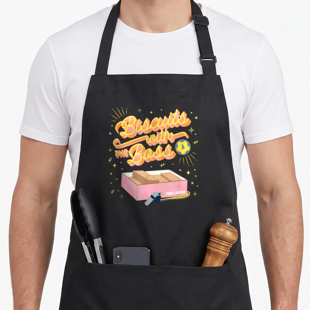 Funny biscuit boss apron - Biscuits with THE Boss text, pink box, whistle, blue cookie, yellow stars — shown on a black cotton canvas apron against a white background