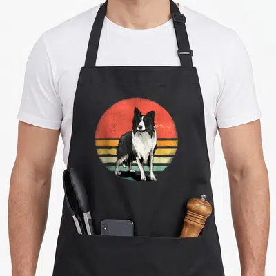 Pet border collie apron - Black and white dog standing in front of a red, yellow, and teal striped retro sun graphic — shown on a black cotton canvas apron against a white background