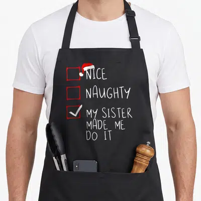 Sister santa claus apron - Nice Naughty My Sister Made Me Do It checklist with red boxes and Santa hat — shown on a black cotton canvas apron against a white background