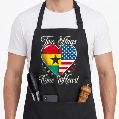 Ghanaian ghana american flag apron - Heart split into Ghana and USA flags with Two Flags One Heart script — shown on a black cotton canvas apron against a white background